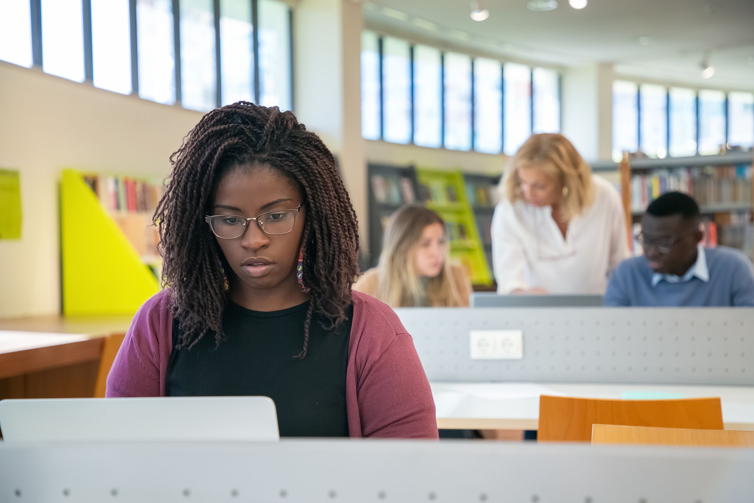 Concentrated African American female student surfing laptop while diverse classmates discussing study task with teacher